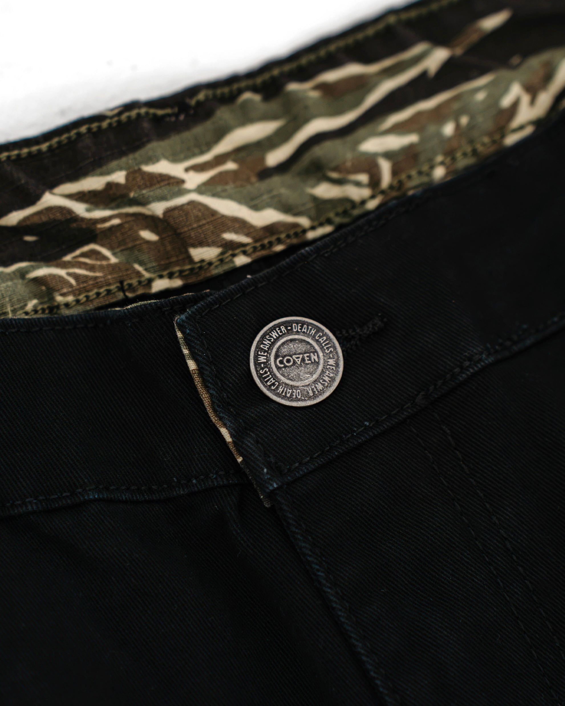 Dead Drop Chino Aeon Tigerstripe in Schwarz, Bunddetail mit COVEN Metallknopf und Tigerstripe-Innenfutter