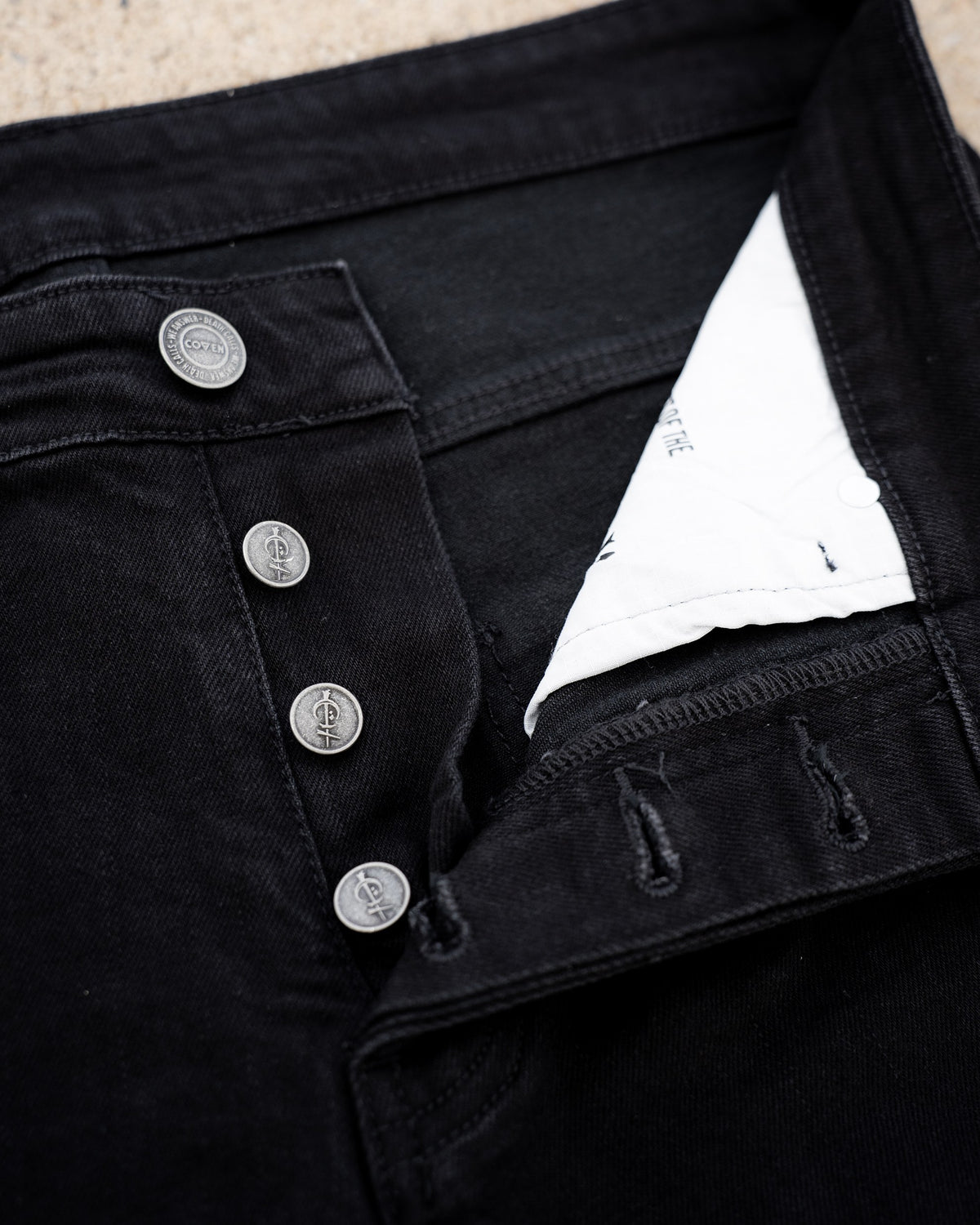 The Wormskin Jeans in Schwarz, Knopfleiste im Makro mit vier geprägten COVEN Knöpfen